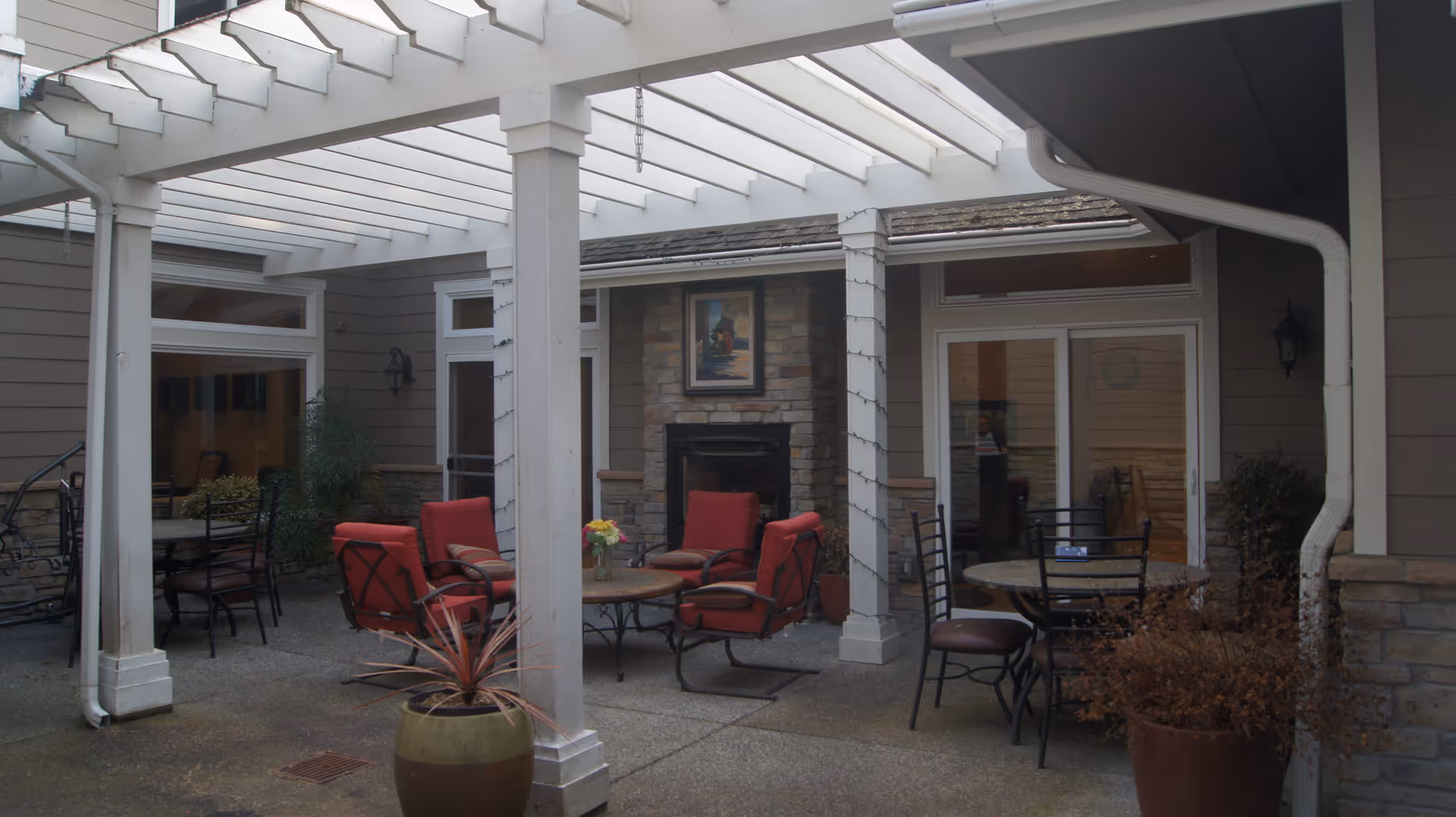 Outdoor patio area with a white pergola overhead, featuring red cushioned chairs around a round table, additional black metal chairs and tables, potted plants, and a stone fireplace with a framed picture above it.