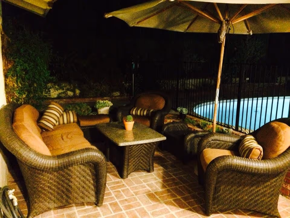 Outdoor patio area at night with wicker furniture including a sofa, two armchairs, a coffee table with a small potted plant, and a large umbrella. There is a swimming pool with a black metal fence in the background and some greenery along the wall.