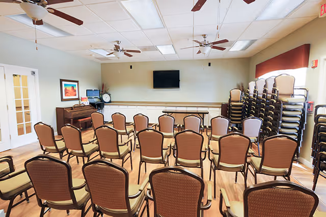 Empty meeting/activity room with rows of chairs facing a wall-mounted TV, a piano, and stacks of extra chairs.