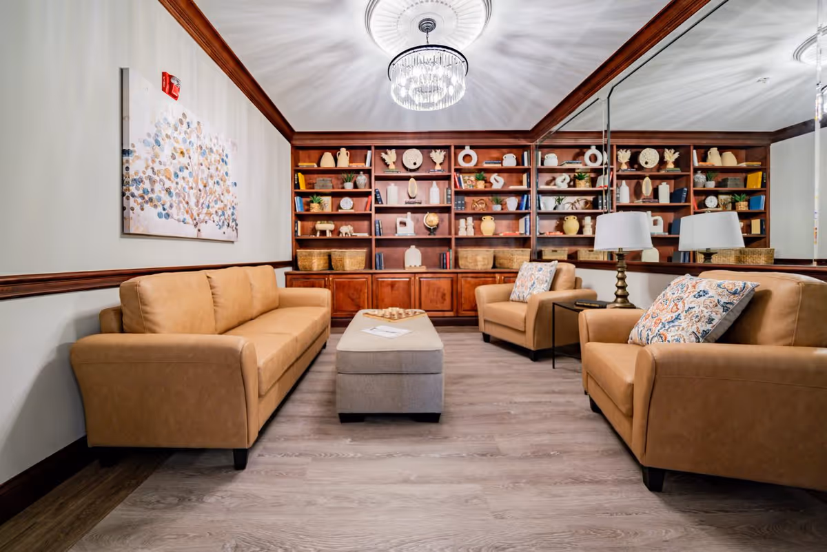 A cozy living room with tan leather sofas and armchairs arranged around a beige ottoman. The room features a large wooden bookshelf filled with decorative items and books, a modern chandelier on the ceiling, and a large mirror on one wall reflecting the space. A colorful abstract painting hangs on the opposite wall.