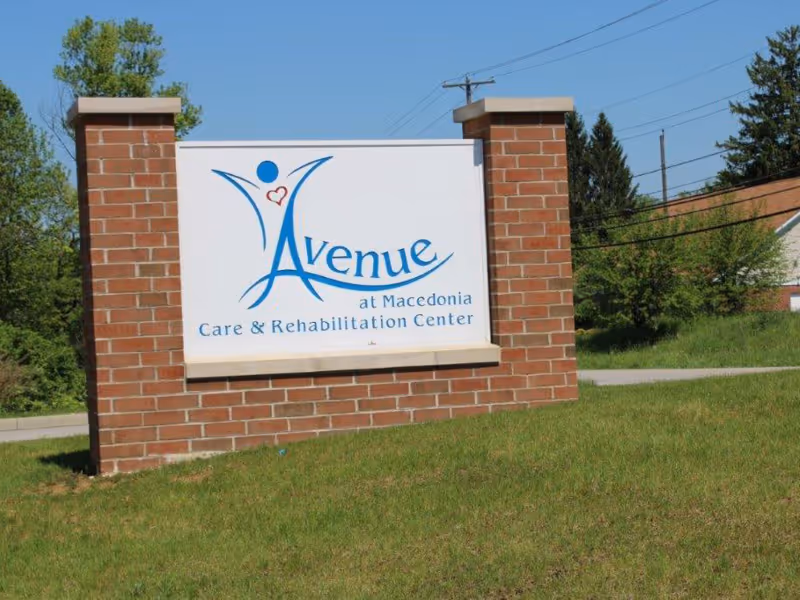 A brick signpost on a grassy area displaying the name 'Avenue at Macedonia Care & Rehabilitation Center' with a blue and red logo featuring a stylized figure with a heart.