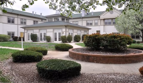 Courtyard with landscaped shrubs and a circular brick planter in front of a low white multi-story senior living building with French doors and a green roof.