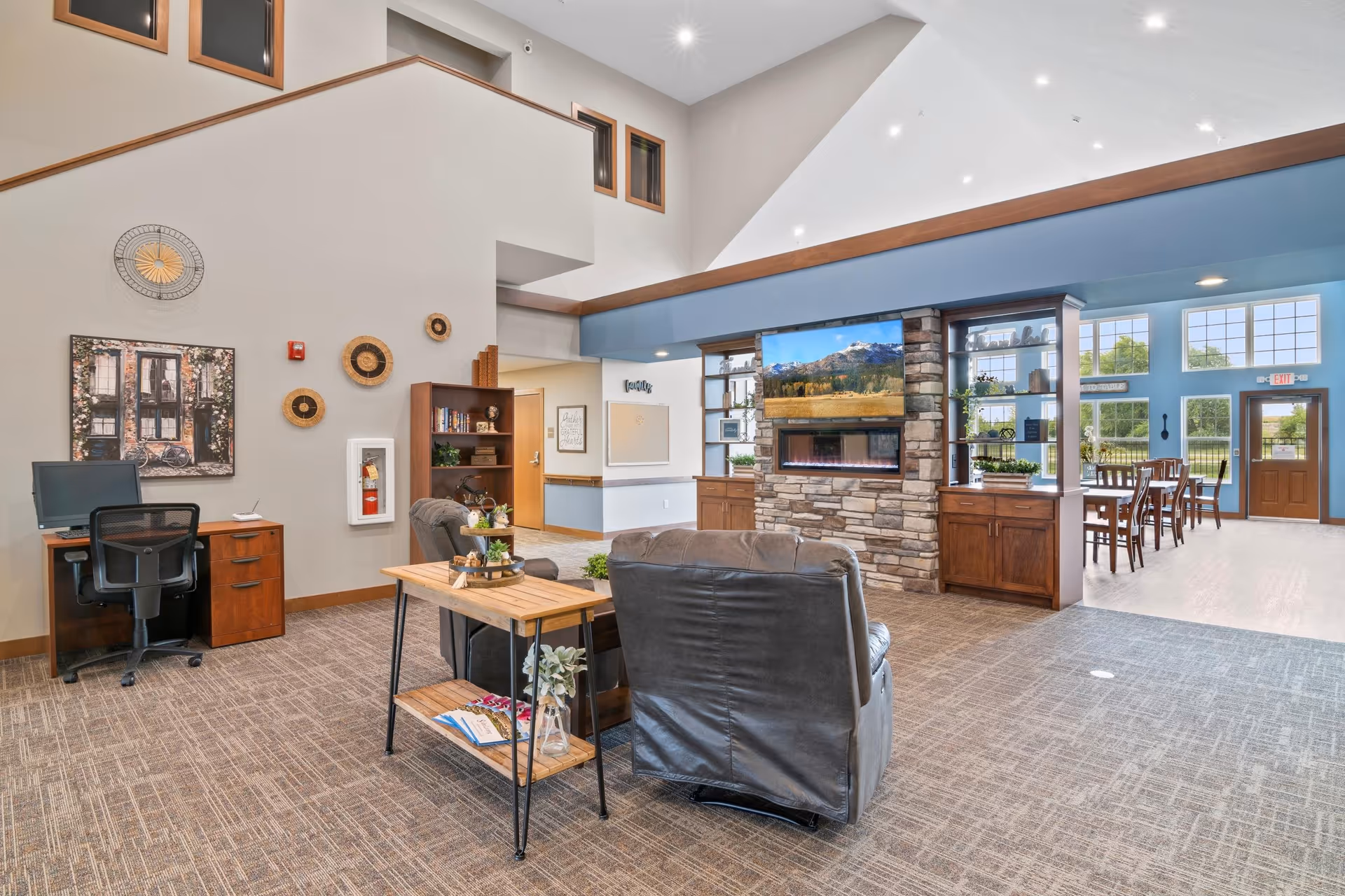 A spacious and well-lit common area in a senior living facility featuring two gray armchairs facing a stone fireplace with a mounted flat-screen TV above it. To the right, there is a dining area with wooden tables and chairs near large windows letting in natural light. On the left side, there is a small office desk with a computer and chair, wall decorations including a clock and woven baskets, and a bookshelf. The room has high ceilings with recessed lighting and a carpeted floor.