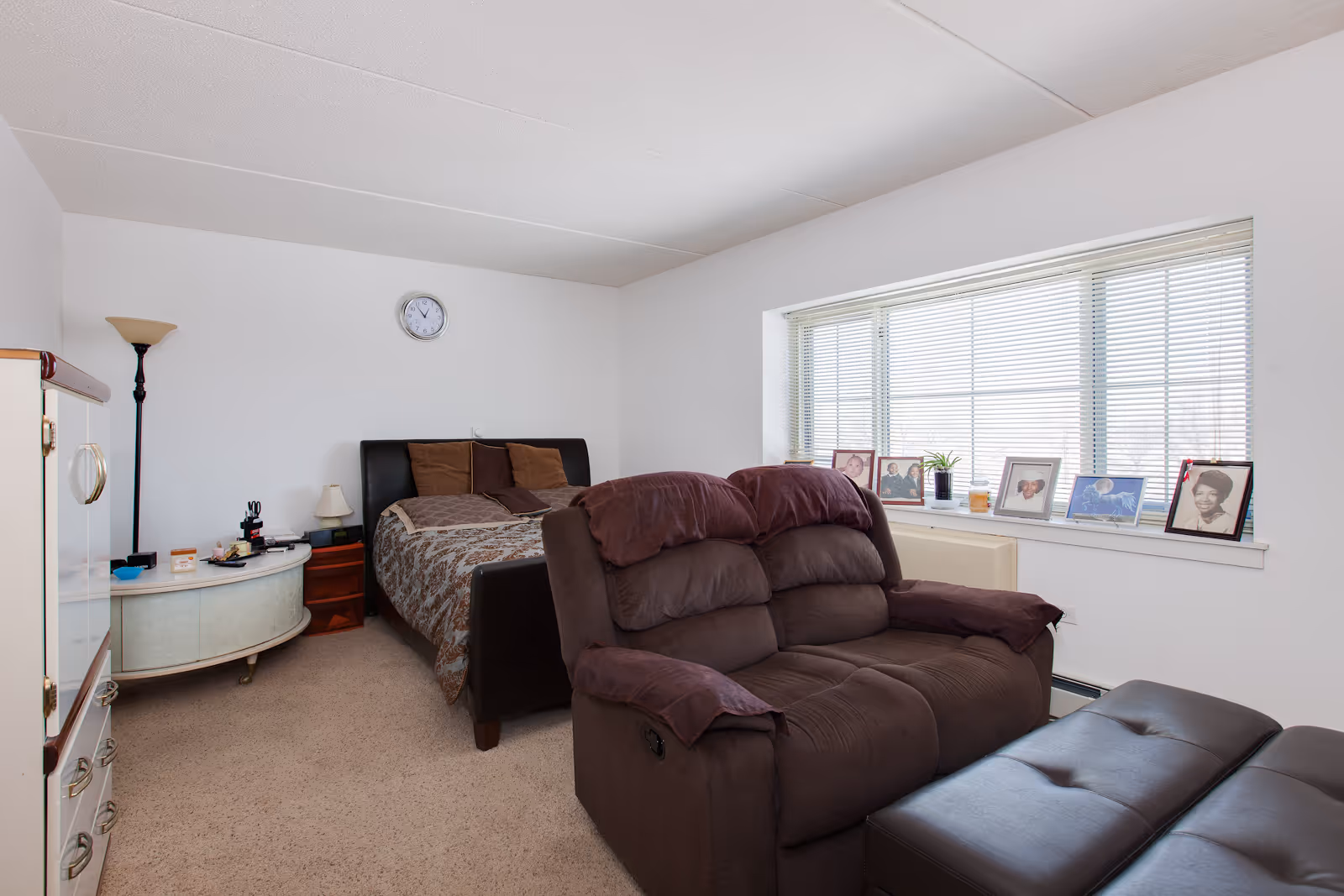 A senior living bedroom featuring a bed with brown and beige bedding, a brown reclining loveseat, and a matching ottoman. The room has a large window with blinds and a windowsill displaying framed photos and a small plant. There is a round side table with a lamp and other small items, a tall floor lamp, and a wall clock above the bed. The walls are white and the carpet is beige.