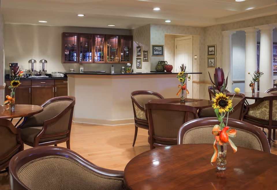 A cozy dining area with round wooden tables and cushioned chairs. Each table has a small vase with a sunflower and decorative ribbons. In the background, there is a counter with glass cabinets above it, containing various decorative items. The room has soft lighting and light-colored walls with framed pictures.