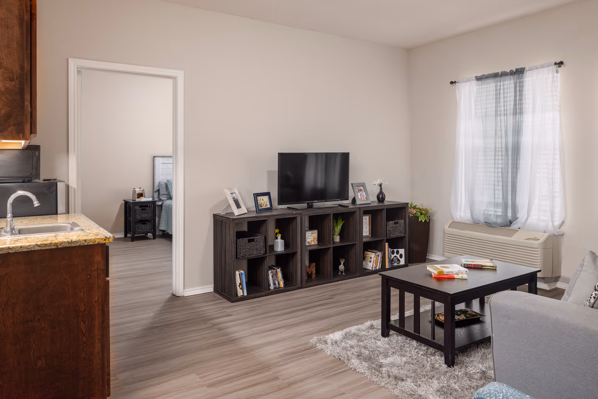A cozy living room area with a dark wood coffee table on a light gray shag rug, a gray sofa, and a dark wood TV stand with a flat-screen TV and decorative items. To the left is a kitchen counter with a sink and granite countertop. A doorway leads to a bedroom with a bed and nightstand. A window with sheer white and gray curtains is on the right wall above an air conditioning unit.