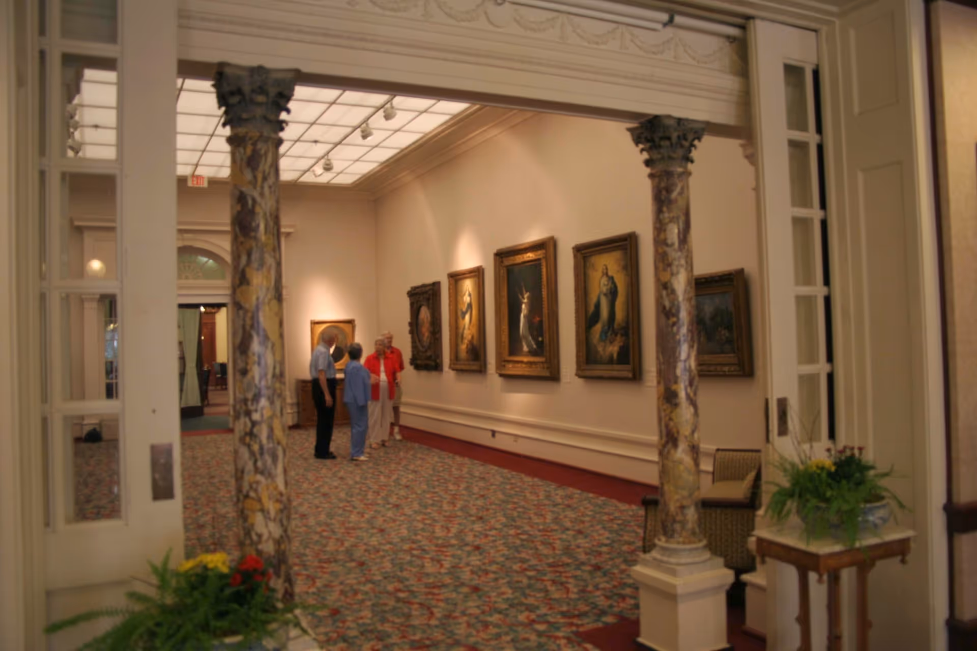 Interior gallery hallway with framed paintings on the wall, two decorative columns, patterned carpet, and a small group of people viewing the art.