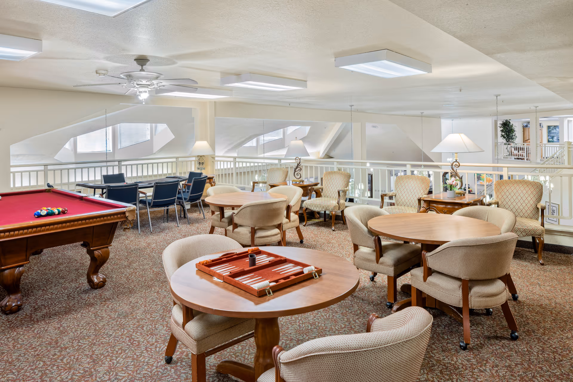 A spacious senior living community game room with a red felt pool table, several round wooden tables with cushioned chairs, a backgammon board on one table, and additional seating areas with armchairs and lamps. The room has a carpeted floor and large windows allowing natural light.