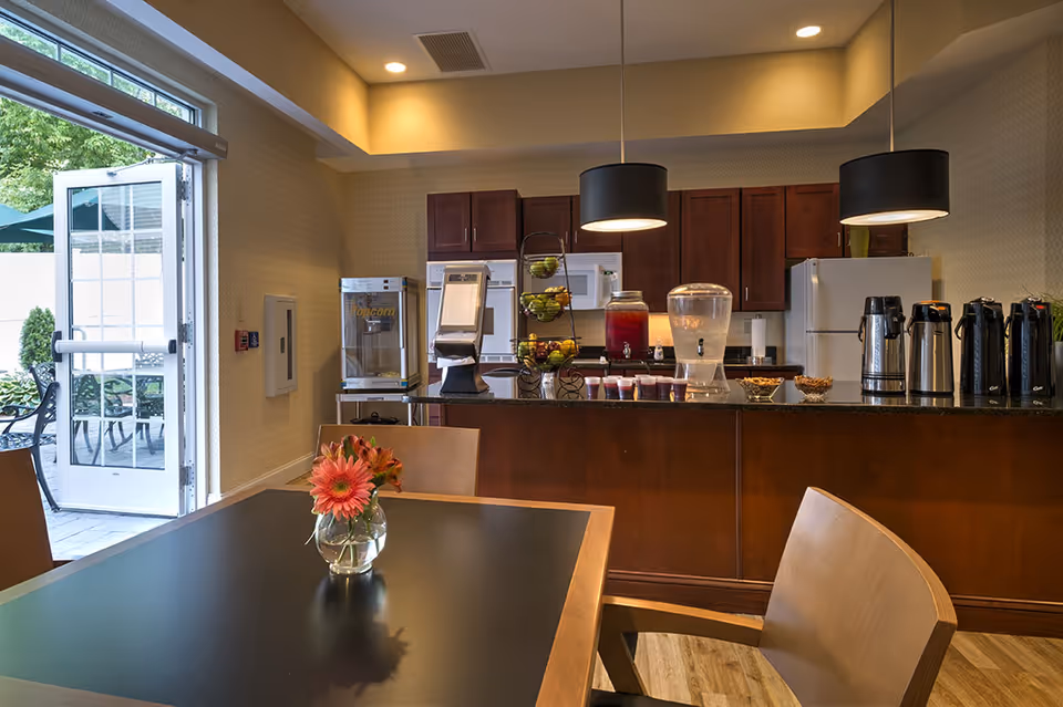 Interior view of a kitchen and dining area in an assisted living facility. The kitchen features dark wood cabinets, a white refrigerator, microwave, and a popcorn machine. On the counter, there are beverage dispensers, coffee urns, bowls of snacks, and a tiered fruit basket. In the foreground, there is a dining table with a small vase holding orange flowers. The room is well-lit with recessed lighting and pendant lights, and a door opens to an outdoor patio area with tables and chairs.