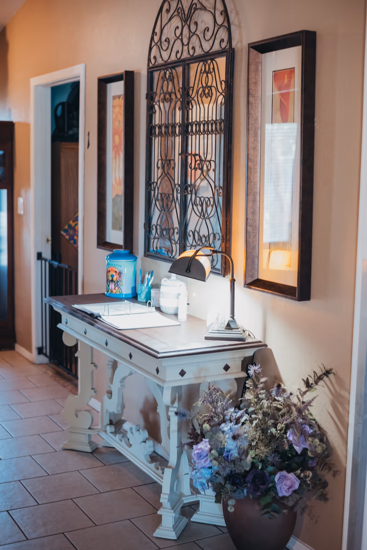 A decorative hallway table with a lamp, a blue container, and some small items on top. Above the table is an ornate wrought iron wall decoration flanked by two framed pictures. A large vase with a floral arrangement sits on the floor next to the table. The setting appears to be a warmly lit interior space with tiled flooring.