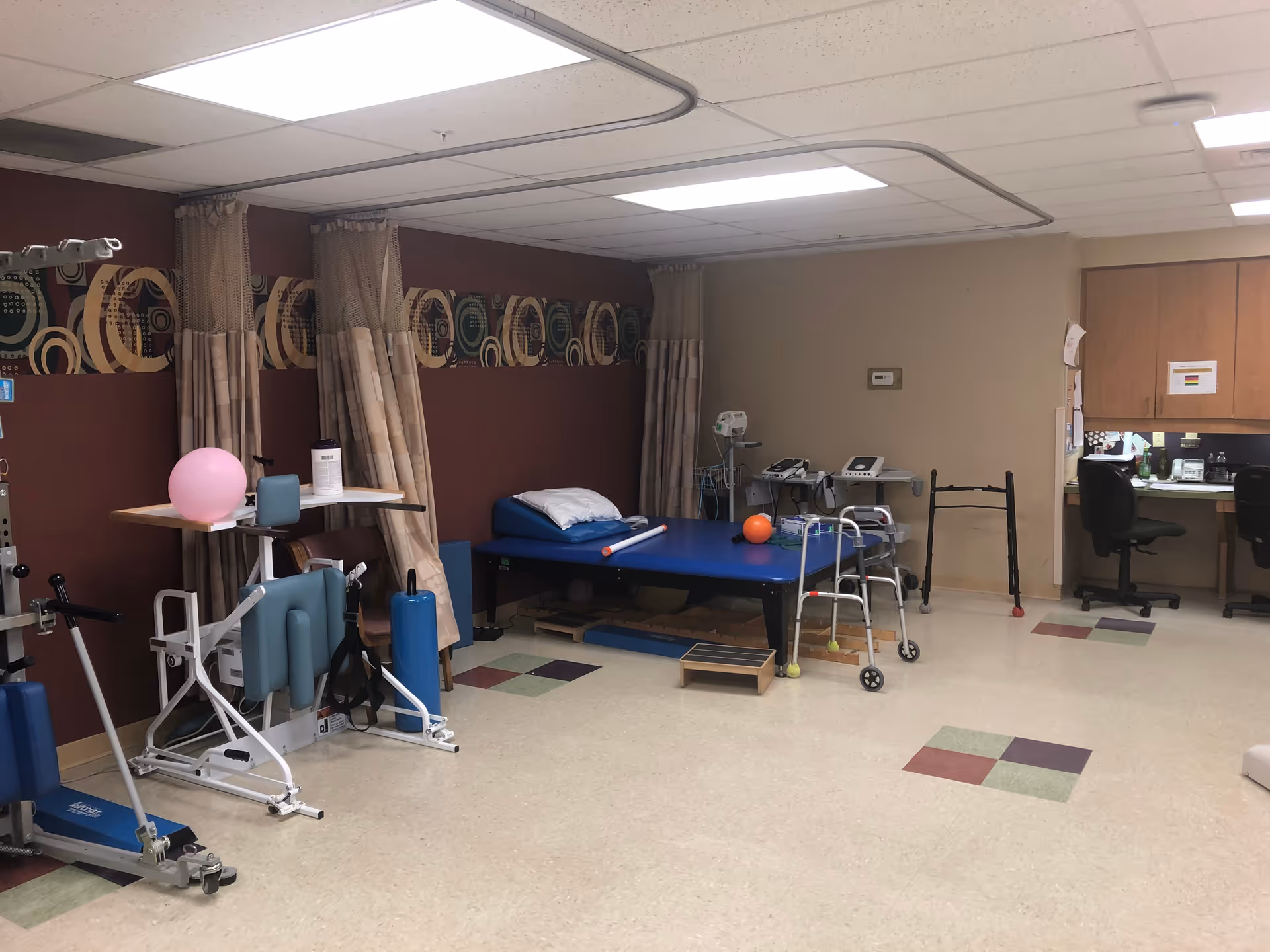 A therapy or rehabilitation room with exercise equipment, a blue therapy table with pillows and an orange ball, a walker, and two office chairs at a desk with cabinets above. The walls are decorated with a patterned border and beige curtains.