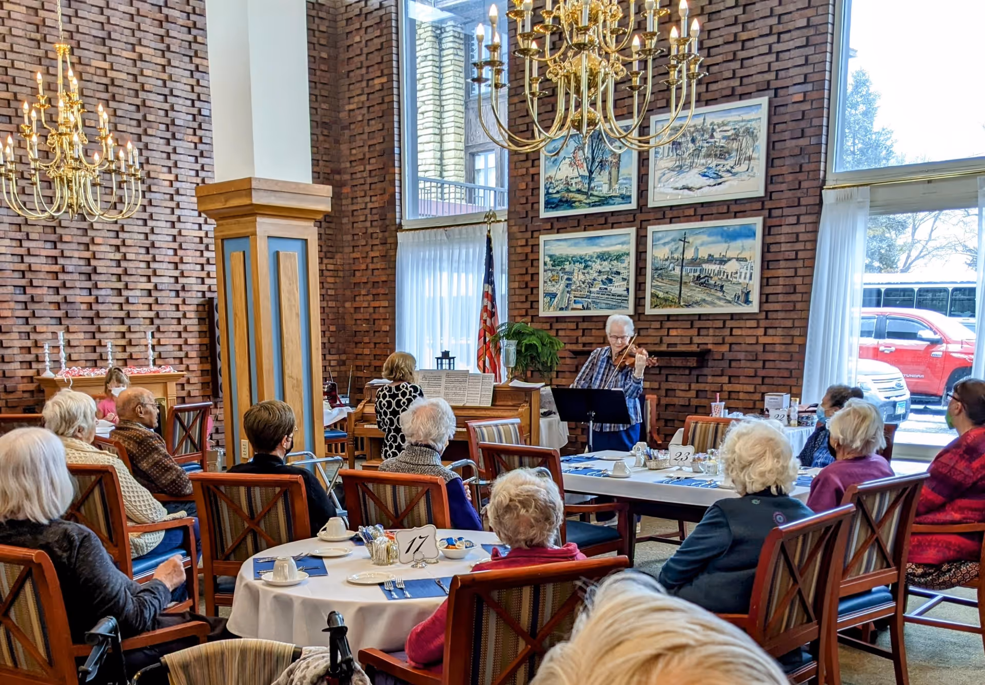 A group of elderly people seated at round tables in a well-lit room with large windows and brick walls, attentively watching a woman playing the violin. The room features chandeliers, framed artwork on the walls, and an American flag near the window.
