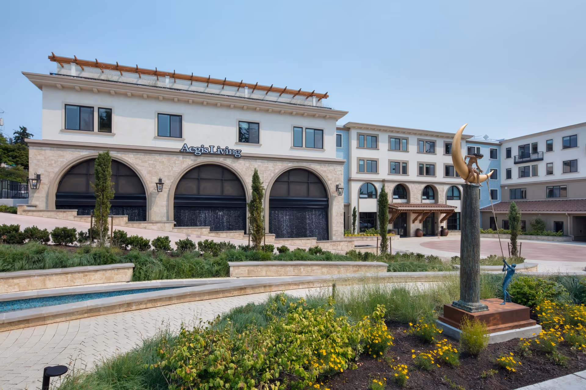 Exterior view of Aegis Living Kirkland Waterfront, showing a multi-story building with arched windows and a landscaped garden in the foreground. There is a decorative sculpture featuring a crescent moon and two figures near the entrance.