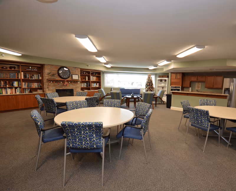 A spacious common area with round tables and patterned chairs arranged for group seating. In the background, there is a fireplace with a clock and bookshelves on either side. A Christmas tree is visible near a large window, and a kitchen area with wooden cabinets and stainless steel appliances is on the right side.