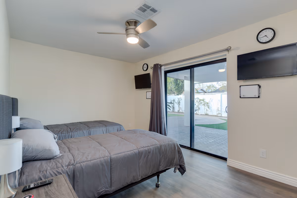 A bedroom with two twin beds covered in gray bedding, a ceiling fan with light, a nightstand with a lamp and remote control, two wall clocks, a wall-mounted TV, and sliding glass doors leading to an outdoor patio area.