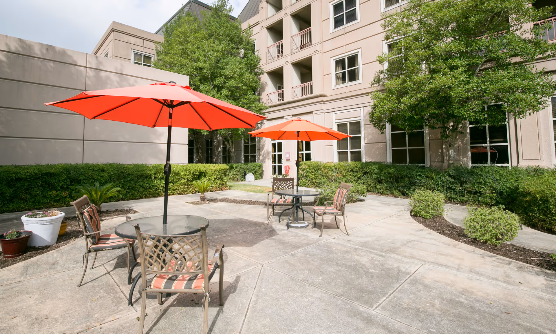 Outdoor patio area at Parklane West Healthcare Center with two round glass tables, each shaded by a large red umbrella. Each table is surrounded by metal chairs with striped cushions. The patio is surrounded by greenery, including bushes and trees, and the building's beige exterior with windows and balconies is visible in the background.