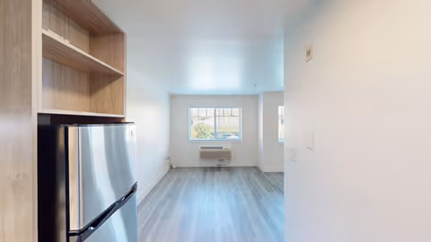 Interior view of a small apartment or studio with light-colored walls and flooring. A stainless steel refrigerator is partially visible on the left, next to wooden shelving. Two windows at the far end let in natural light, and there is an air conditioning unit mounted below the windows.