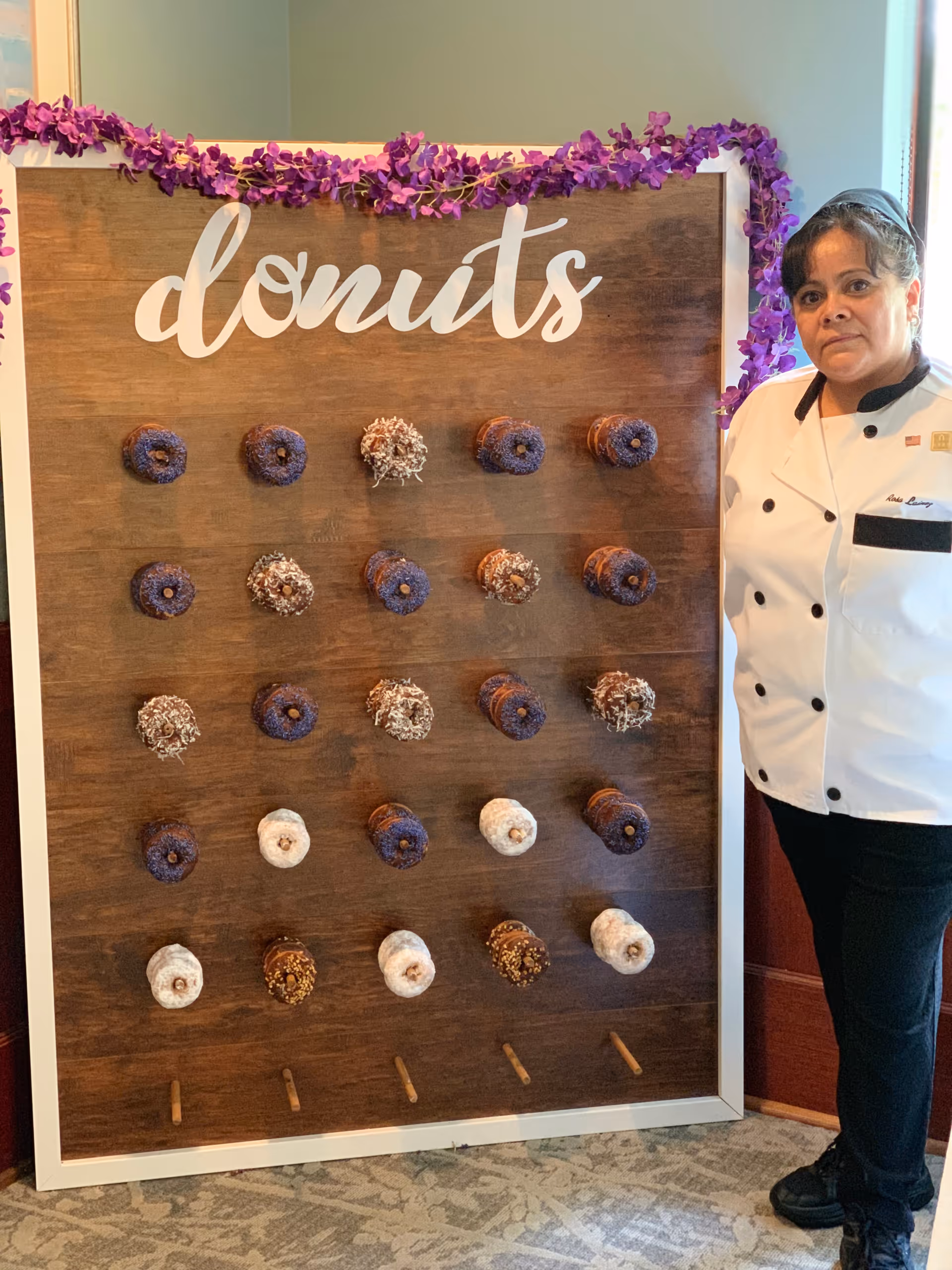 A woman in a white chef's coat stands next to a wooden donut wall decorated with purple flowers. The wall has the word 'donuts' written at the top and holds multiple donuts with various toppings arranged in rows.