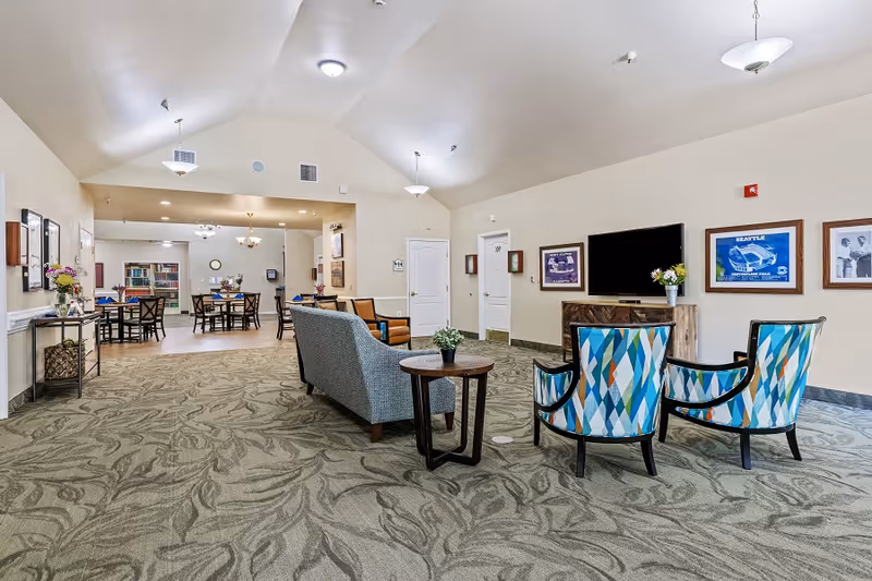 A spacious senior living common area with patterned carpet, a sofa, two colorful armchairs, a round side table with a small plant, and a flat-screen TV mounted on the wall. The room has high vaulted ceilings with hanging light fixtures. In the background, there is a dining area with several tables and chairs, decorated with flowers and blue napkins.