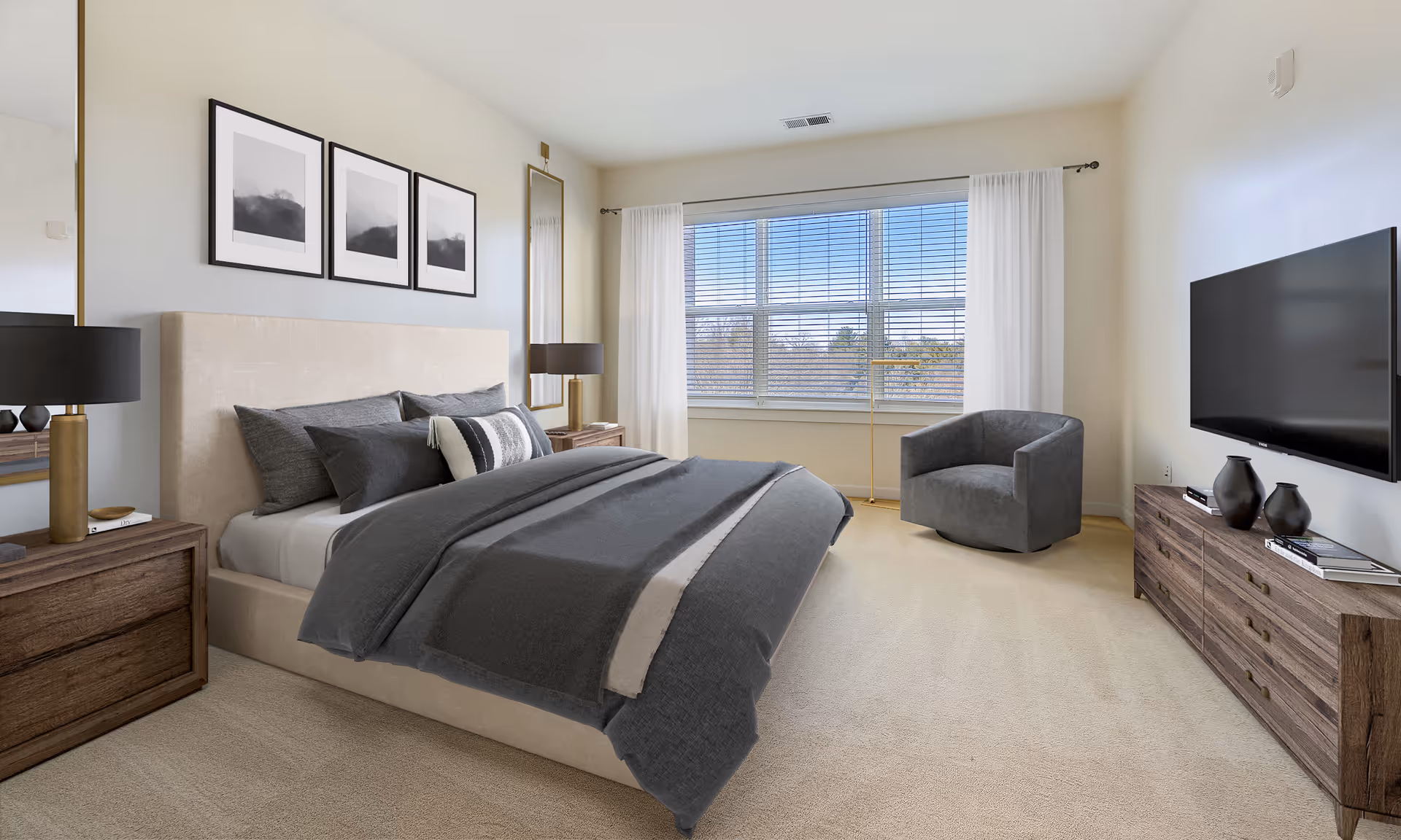 A bright bedroom with a large beige upholstered bed covered with gray bedding and pillows. There are two wooden nightstands with black lamps on either side of the bed. Above the bed are three framed abstract art pieces. A tall mirror is positioned next to the bed. A large window with white curtains and blinds lets in natural light. Across from the bed is a wooden dresser with decorative vases and books, and a flat-screen TV mounted on the wall. A gray upholstered armchair sits near the window.