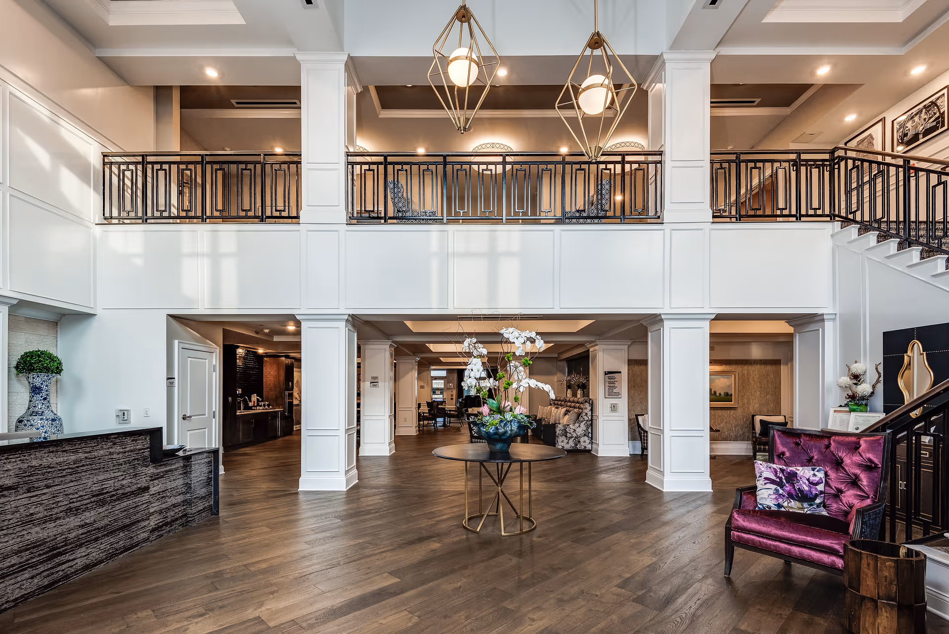 Spacious and elegant senior living facility lobby with high ceilings, a central round table adorned with a floral arrangement, a purple upholstered chair with a floral cushion, a reception desk on the left, and a staircase on the right leading to an upper balcony with decorative railings and seating.