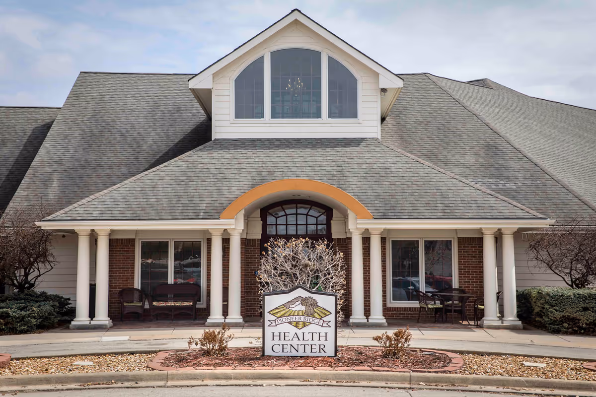 Front exterior view of Pioneer Ridge Health Center building with a peaked roof, white columns, and a sign in front that reads 'Pioneer Ridge Health Center'. There are chairs and tables on the porch area on either side of the entrance.