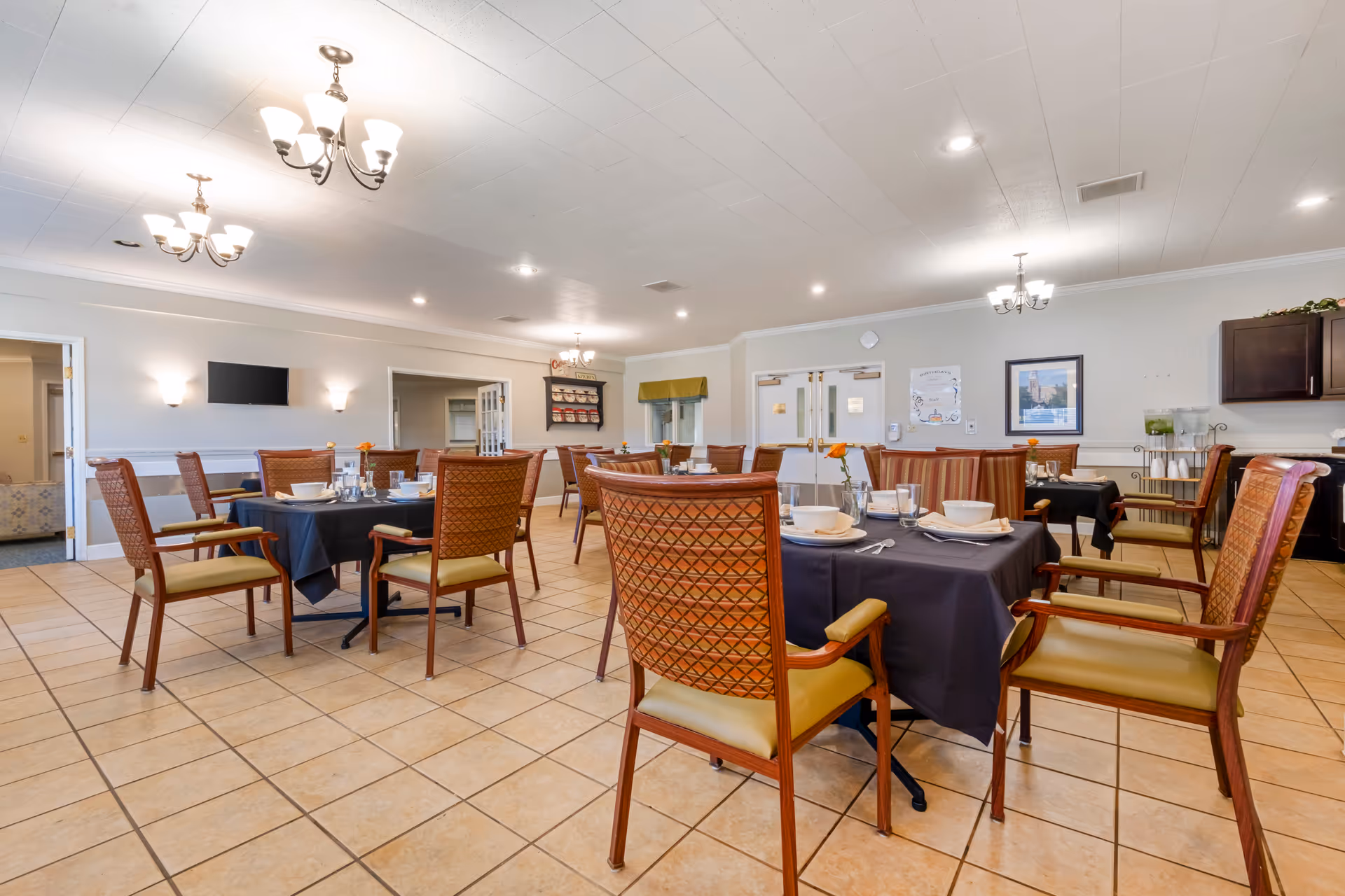 A spacious dining room with several tables covered in dark tablecloths, each set with bowls, plates, cups, and silverware. The room features tiled floors, wooden chairs with cushioned seats, and multiple chandeliers hanging from the ceiling. There are framed pictures and a water dispenser on the far wall, and double doors in the background.