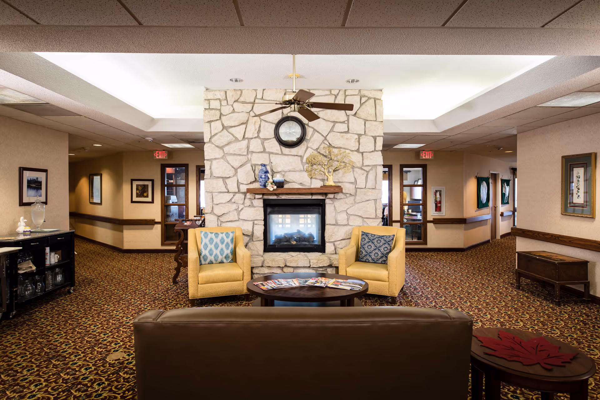 A cozy common area in Twin Oaks Independent Living of Lansing featuring a stone fireplace with a clock above it, two yellow armchairs with patterned cushions, a round wooden coffee table with magazines, and a brown leather sofa. The room has patterned carpet, framed artwork on the walls, and ceiling lights with a ceiling fan above the fireplace.