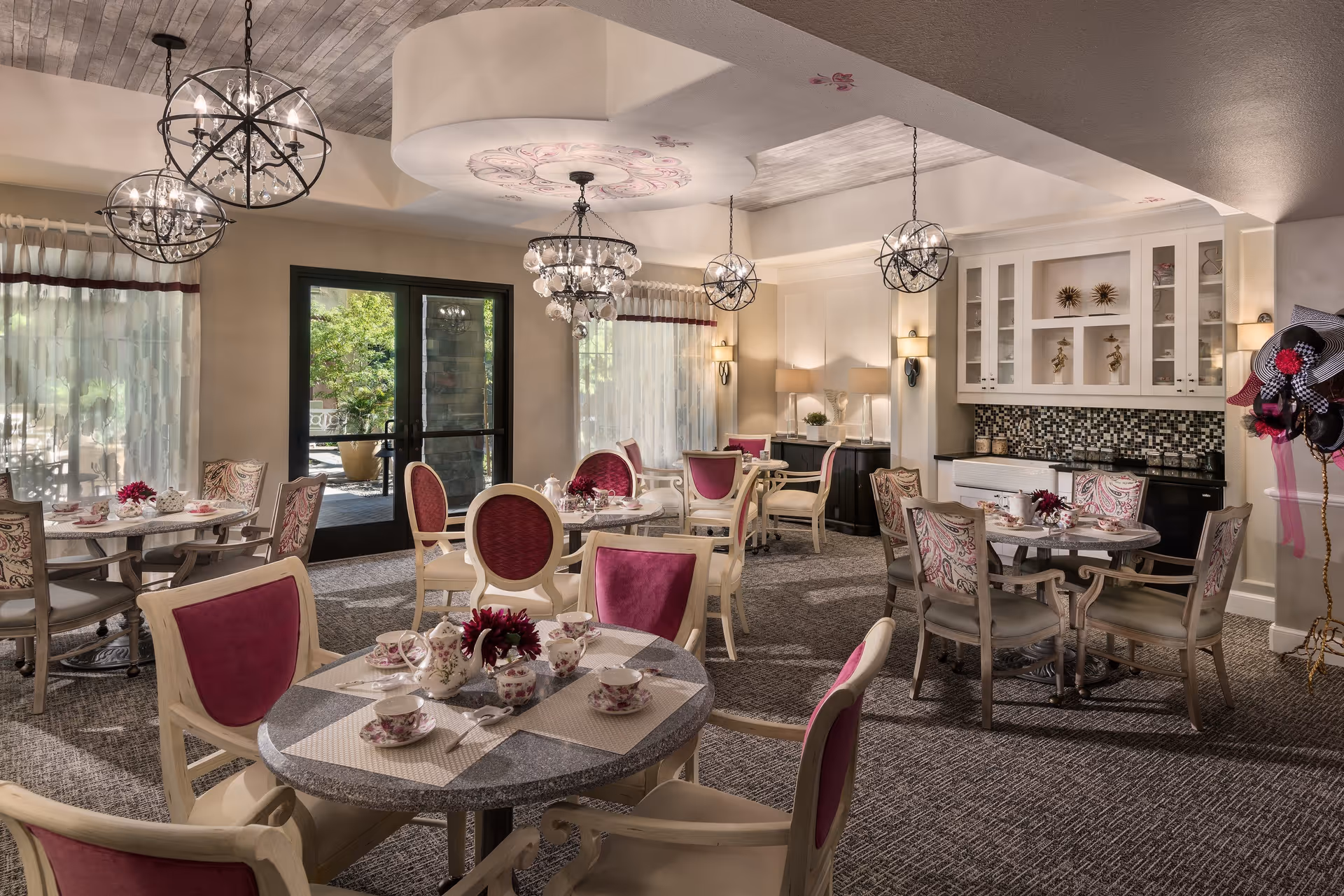 A cozy dining room with round tables set with floral-patterned tea sets and pink flowers. The chairs have pink and patterned upholstery. The room features elegant chandeliers, large windows with sheer curtains, and a door leading outside. There is a built-in cabinet with decorative items and a mosaic tile backsplash on one wall.