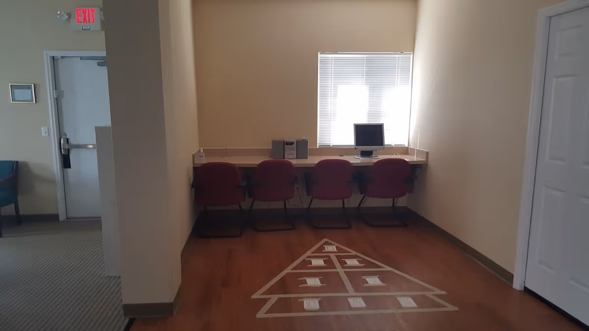 A small room with four red chairs lined up against a wall-mounted desk. On the desk, there is a computer monitor and a stereo system. The floor has a shuffleboard court marked on it. There is a window with blinds above the desk, a door on the right, and an exit door visible in the adjacent room to the left.