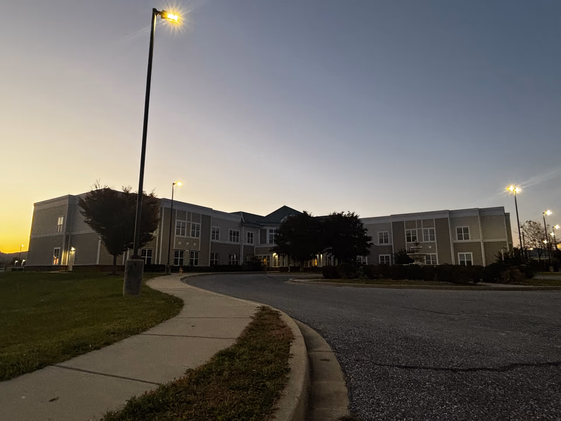 Exterior view of a two-story assisted living facility building at dusk with street lamps lit along a curved driveway and sidewalk. The sky is clear with a gradient from light near the horizon to darker blue above. Trees and shrubs are visible around the building.