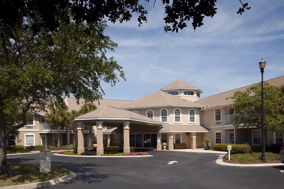 Exterior view of Allegro Senior Living facility in Fleming Island, FL, showing a beige two-story building with a covered entrance, surrounded by trees and landscaping under a clear blue sky.