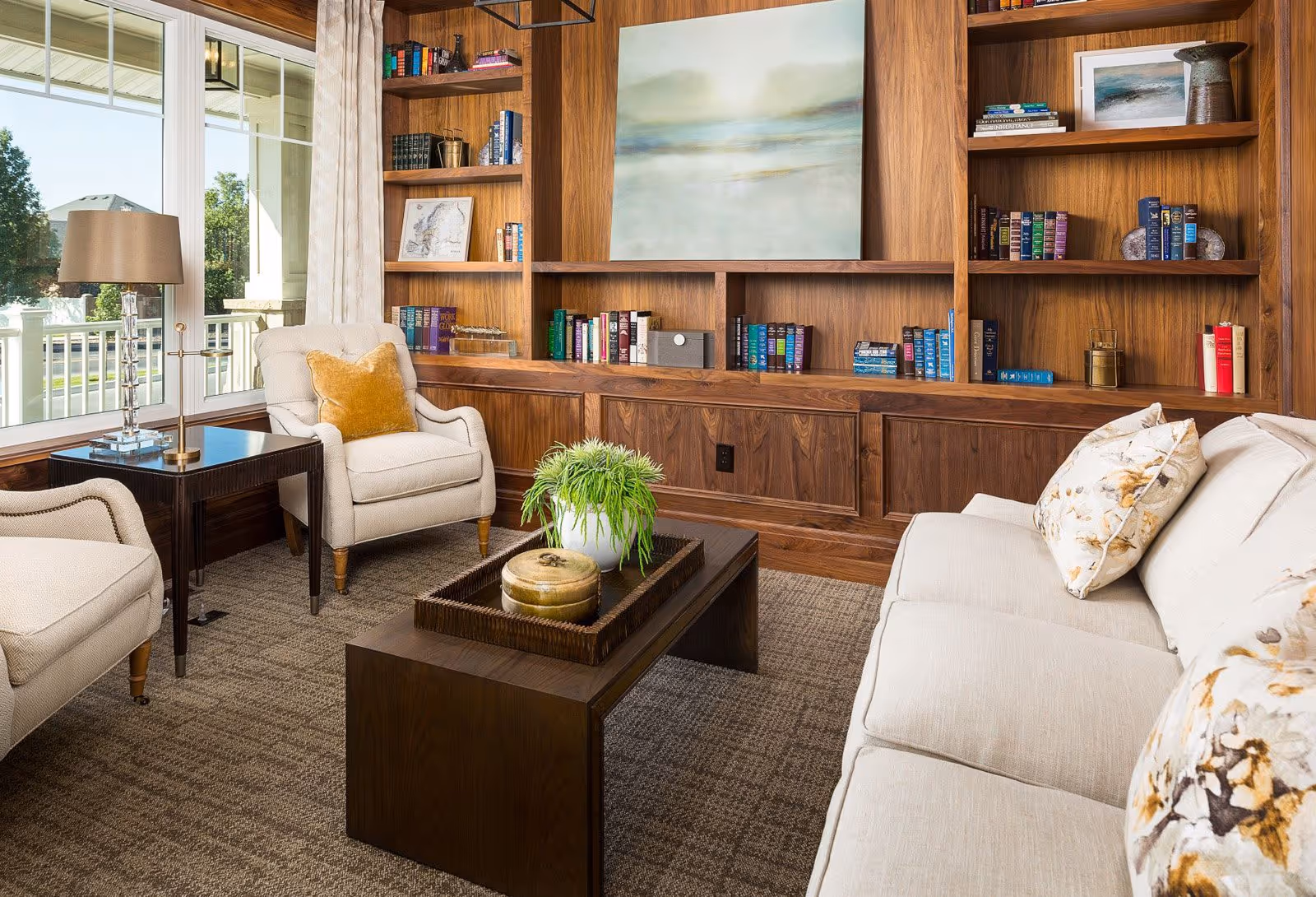 A cozy living room with a beige sofa adorned with floral pillows, two beige armchairs with a mustard yellow pillow on one, a dark wooden coffee table with a green plant and decorative items, and built-in wooden bookshelves filled with books and decor. Large windows let in natural light and show an outdoor view.