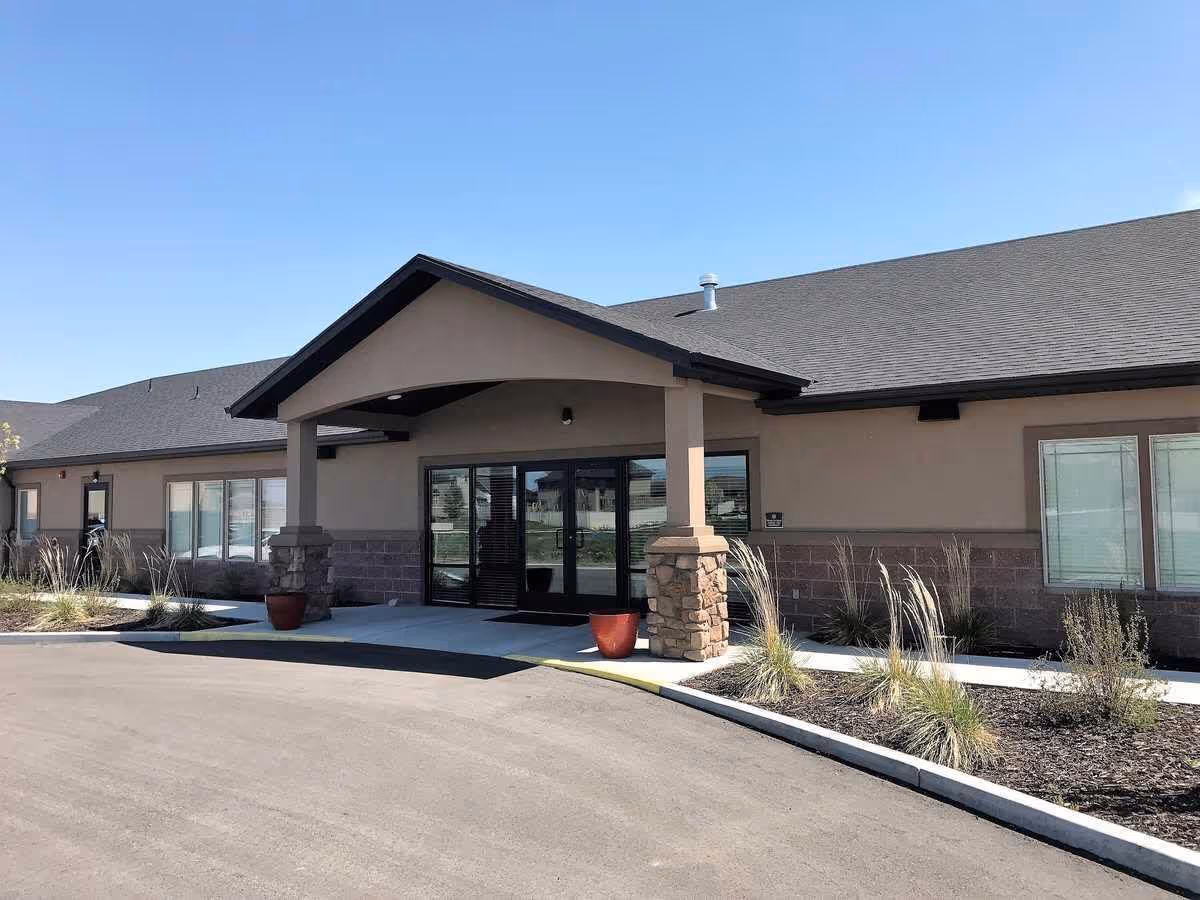 Exterior front entrance of a single-story building with a covered porch supported by stone pillars, large glass double doors, and windows on either side. There are landscaped areas with plants and a paved driveway in front under a clear blue sky.