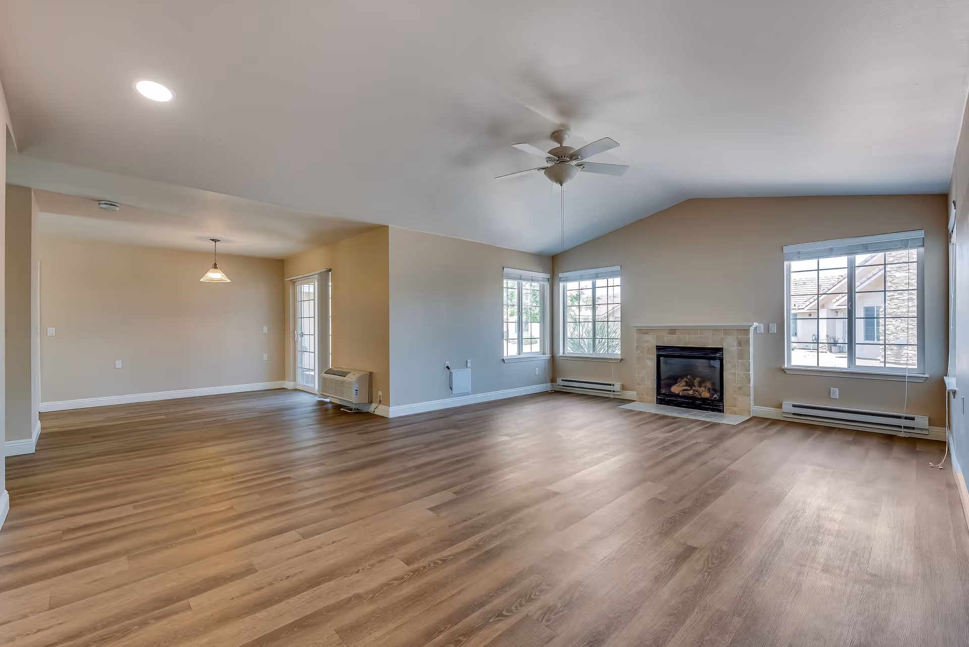 Spacious empty living room area with light wood flooring, beige walls, a ceiling fan, and a fireplace. The room has multiple windows letting in natural light and an adjacent dining area with a hanging light fixture and glass door.