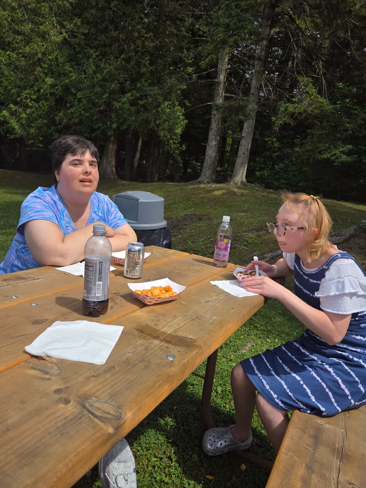 Two women sitting at a wooden picnic table outdoors, eating snacks and drinks. One woman is wearing a blue striped dress and glasses, and the other is wearing a blue patterned shirt. There are trees and grass in the background.