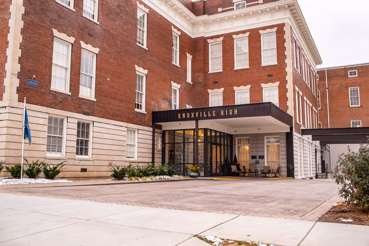 Exterior view of Knoxville High Apartments, a multi-story brick building with large windows and a covered entrance. There are some shrubs and a flagpole near the entrance, and the building name is displayed above the entrance.