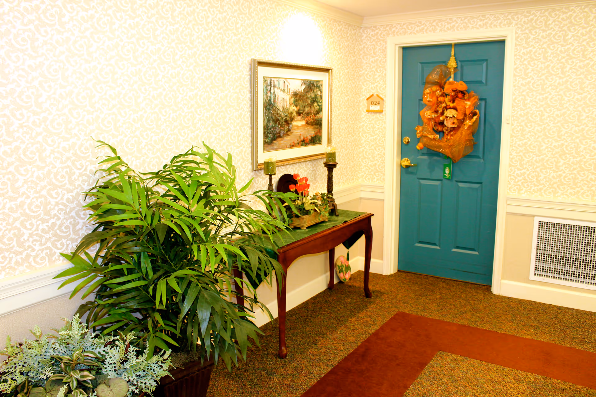 A hallway corner in a senior living facility with a teal door decorated with an orange wreath. Next to the door is a wooden table with decorative items including candles and a small plant. There is a large potted plant on the left side and a framed painting hanging on the wallpapered wall.