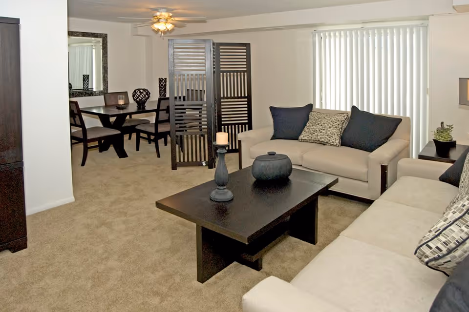 A living room area with beige sofas adorned with black and patterned cushions, a dark wooden coffee table with decorative items, and a dining area in the background separated by a dark wooden room divider. The dining area has a dark wooden table with four chairs and a large mirror on the wall. Vertical blinds cover the window, and the room is carpeted in beige.