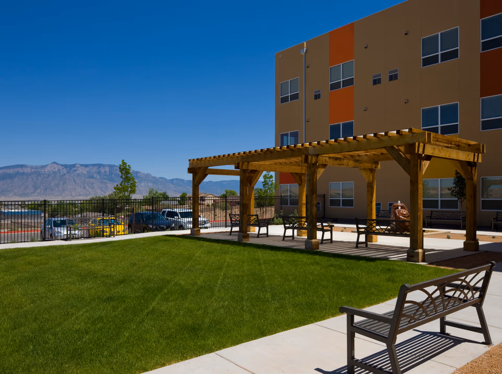 Outdoor seating area with a wooden pergola and several benches on a concrete patio next to a green lawn. In the background, there is a multi-story building with orange and beige walls and multiple windows. Mountains and a clear blue sky are visible in the distance.