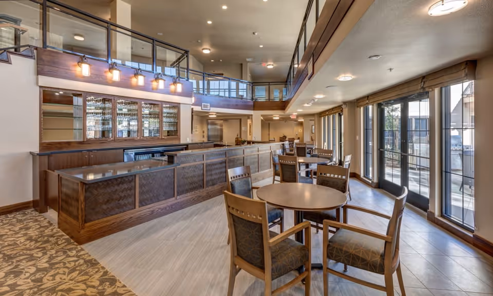 A spacious and well-lit common area with round tables and chairs arranged for seating. The room features large windows and glass doors on one side allowing natural light to enter. There is a wooden bar counter with glass shelves and hanging lights above it. The ceiling has recessed lighting and the floor is a combination of carpet and tile.