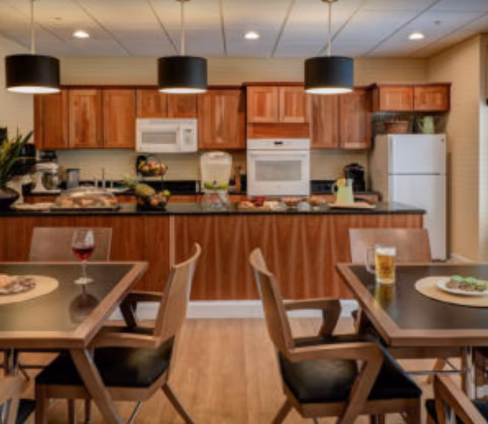 A kitchen and dining area with wooden cabinets, a white microwave, oven, and refrigerator. The kitchen counter has various items including a fruit basket, a water dispenser, and some food. In the foreground, there are two dining tables with wooden chairs, each table having a drink and a plate of food. Three black pendant lights hang from the ceiling above the kitchen counter.