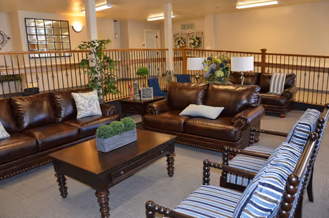 A cozy living room area in a retirement community featuring two brown leather sofas, a wooden coffee table with a small green plant centerpiece, several striped cushioned chairs, decorative pillows, table lamps, and indoor plants. The space has a railing overlooking a lower level and neutral-colored walls with soft lighting.