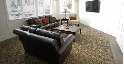 Bright sitting area with leather sofas, an armchair, coffee table, patterned carpet and a wall-mounted TV by large windows.