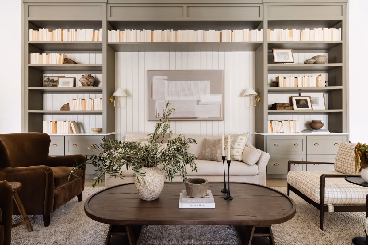 A cozy living room with a beige sofa, a brown armchair, and a plaid armchair arranged around a dark wooden coffee table. The coffee table has a large vase with green foliage, two candlesticks, and some books. Behind the sofa is a built-in bookshelf filled with books and decorative items, and a piece of abstract art hangs on the wall. The room has a neutral color palette with soft lighting.