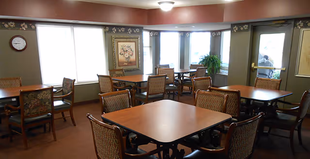 A dining room with multiple square wooden tables and cushioned chairs arranged around them. The room has large windows letting in natural light, a wall clock, a framed floral painting, and a potted plant near the windows. The walls are painted in muted tones with a floral border near the ceiling.
