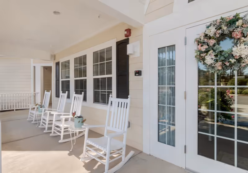 Covered porch area with a row of white rocking chairs and small tables, each table holding a small decorative watering can with flowers. The porch is attached to a building with beige siding, white trim, black shutters, and a glass door adorned with a floral wreath.
