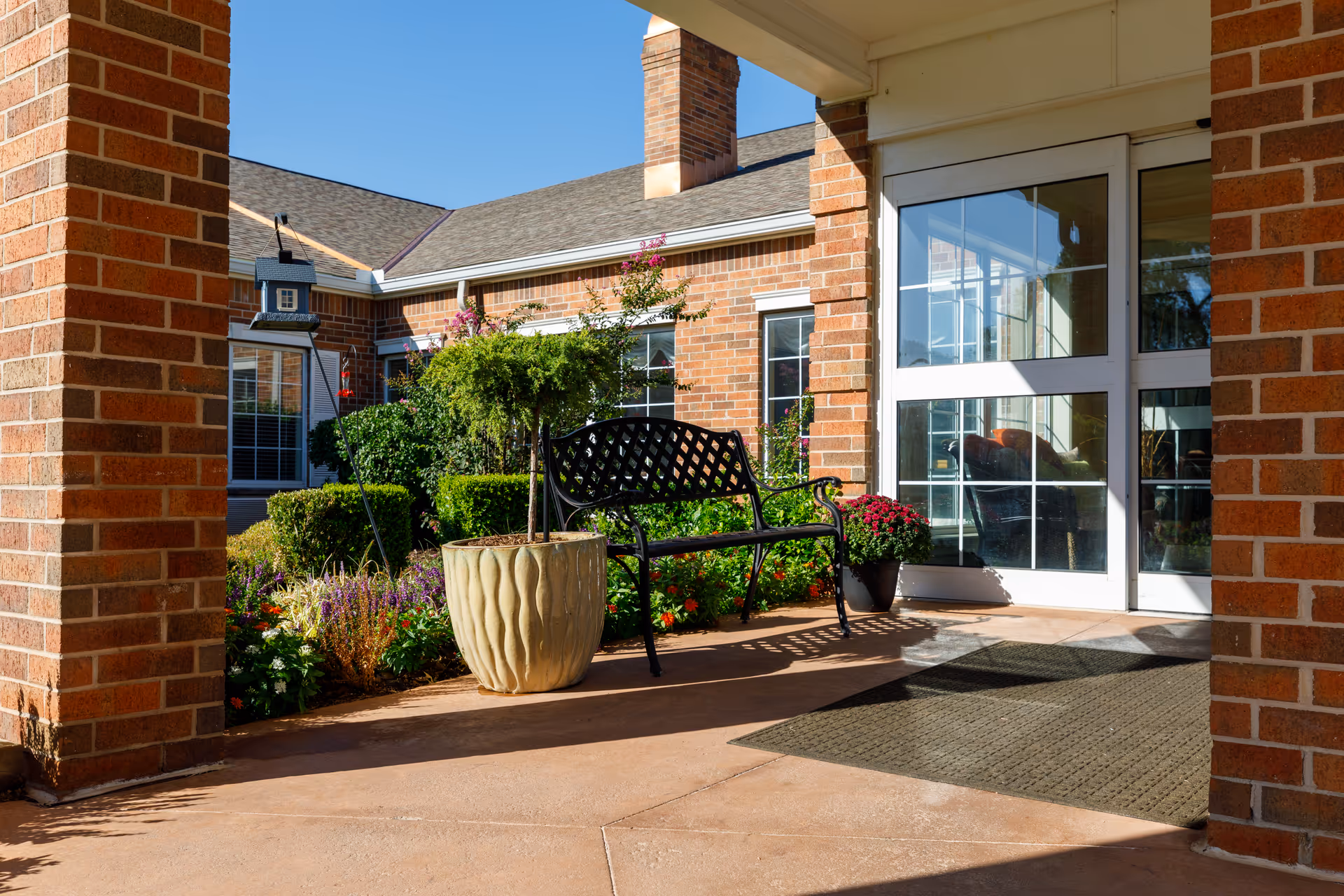Outdoor entrance area of a brick building with a black metal bench, large potted plants, colorful flowers, and glass double doors under a covered porch.
