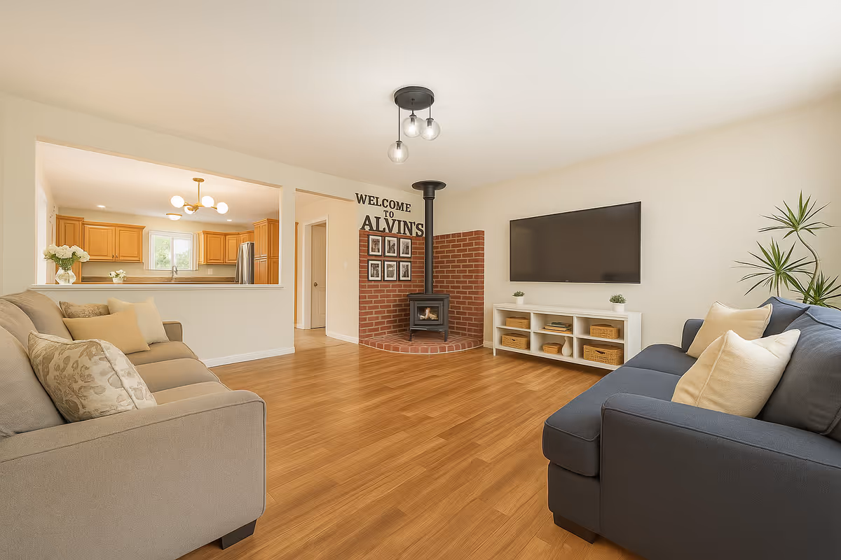A bright and spacious living room with two sofas facing each other, a wall-mounted flat-screen TV above a white media console with baskets, a brick fireplace with a black wood stove, and a large plant in the corner. The room has light wood flooring and cream-colored walls. Through a large pass-through window, a kitchen with wooden cabinets and modern lighting is visible. A wall near the fireplace has a welcome sign that reads 'WELCOME TO ALVIN'S' with framed pictures underneath.