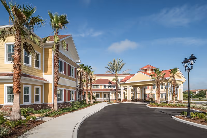 Exterior view of Buffalo Crossings Assisted Living facility featuring a yellow two-story building with white trim, palm trees lining the driveway, a covered entrance with columns, and a clear blue sky.